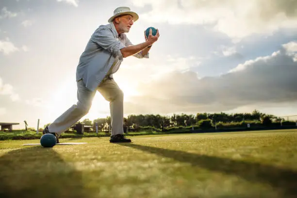 Low angle view of a senior man in position to throw a boule. Old man playing boules in a lawn with sun in the background.