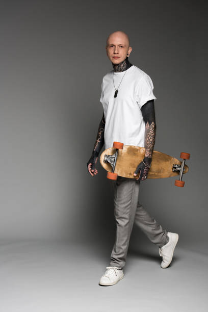 istock handsome tattooed man in white t-shirt walking with longboard and looking at camera on grey 1126086826