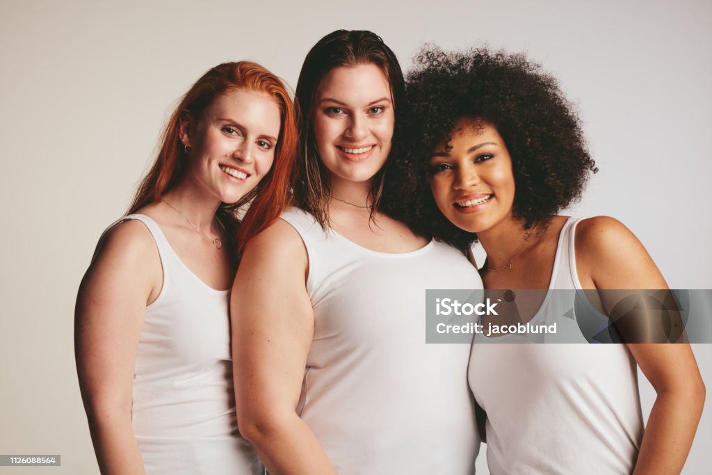 Group of different size women in white tank top Group of women of different size dressed in white tank top standing together. Three diverse women looking at camera against white background. Women Stock Photo
