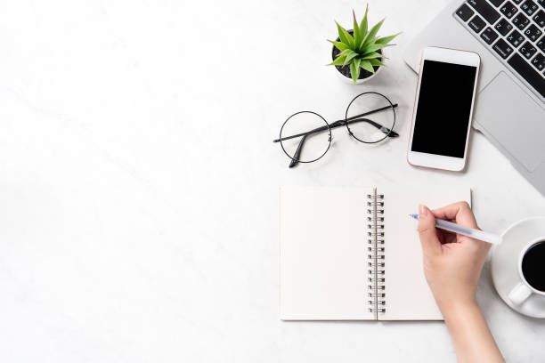 A woman is writing notes isolated on stylized marble office working desk with smartphone, laptop, glasses and coffee, mock up, topview, flatlay, copyspace, closeup