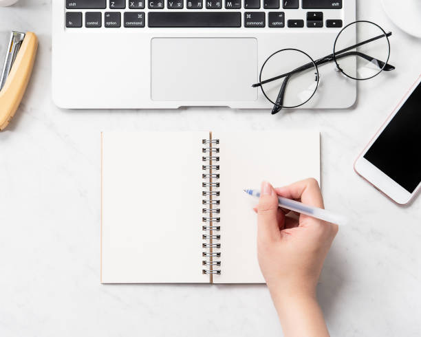 istock A woman is writing notes isolated on stylized marble office working desk with smartphone, laptop, glasses and coffee, mock up, topview, flatlay, copyspace, closeup 1126091610