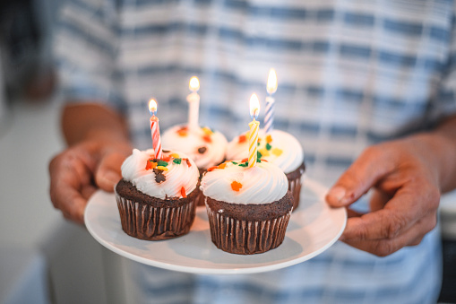 Midsection of senior man holding plate with lit candles on birthday cupcakes. Focus is on delicious dessert for party. Elderly male is at home.