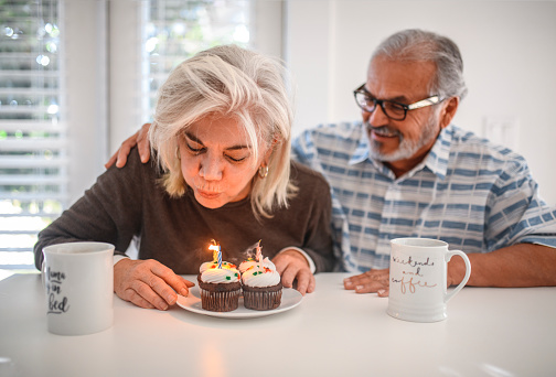 Elderly couple with lit candles on cupcakes enjoying party. Happy senior woman celebrating her birthday with man at home. They are sitting at table. Woman is blowing candles.