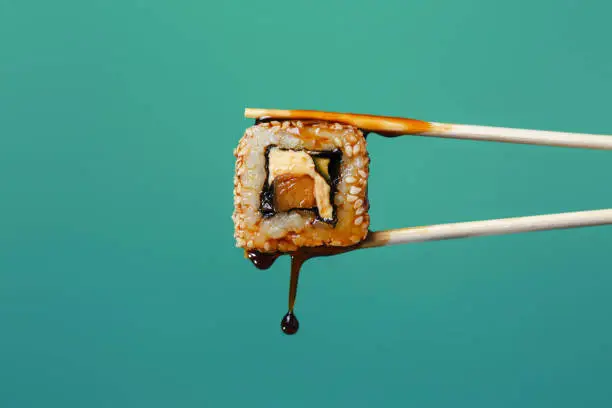 Photo of succulent roll between chopsticks on a colored background, drops of soy sauce dripping from sushi, food background, Japanese cuisine