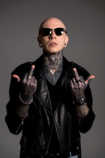 bald tattooed man in leather jacket and sunglasses showing middle fingers isolated on grey