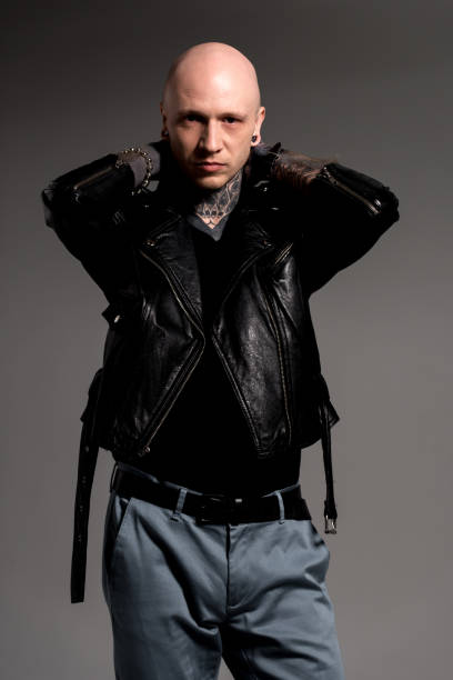 istock stylish tattooed man in leather jacket standing with hands behind head and looking at camera isolated on grey 1126105617