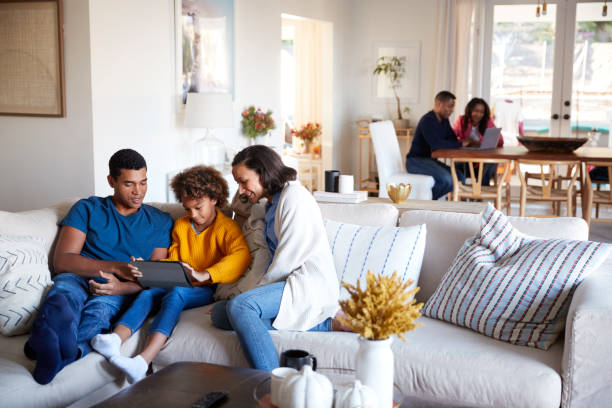 istock Young parents sitting on sofa with their daughter using tablet computer in open plan living room, grandparents sitting at a table in the background 1126136382