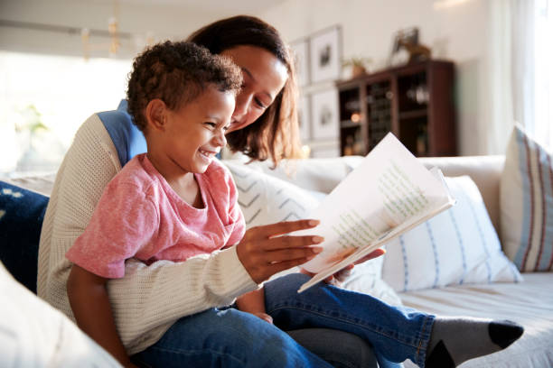 gros plan de jeune maman assise sur un canapé dans le salon, lisant un livre avec son enfant en bas âge, qui est assis sur son genou, vue latérale - family reading african descent book photos et images de collection