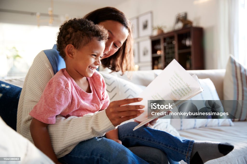 Close up of young mother sitting on a sofa in the living room reading a book with her toddler son, who is sitting on her knee, side view Reading Stock Photo