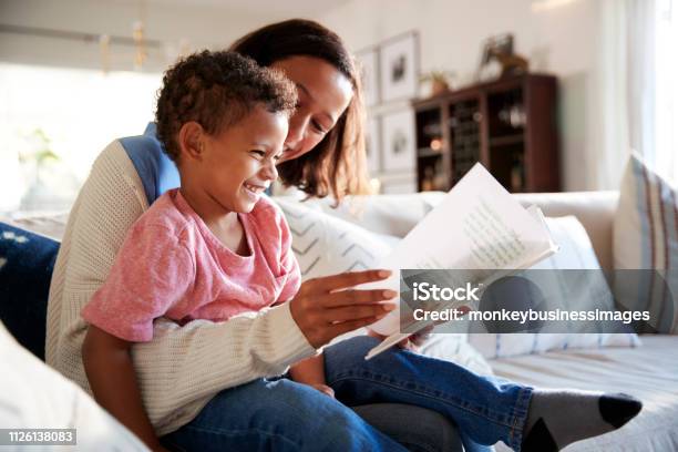 Close Up Of Young Mother Sitting On A Sofa In The Living Room Reading A Book With Her Toddler Son Who Is Sitting On Her Knee Side View Stock Photo - Download Image Now