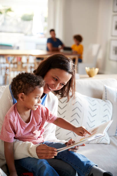 gros plan de jeune maman assise sur un canapé dans le salon, lisant un livre avec son enfant en bas âge, assis sur son genou, le père et la fille assise à une table dans le fond, vertical - family reading african descent book photos et images de collection