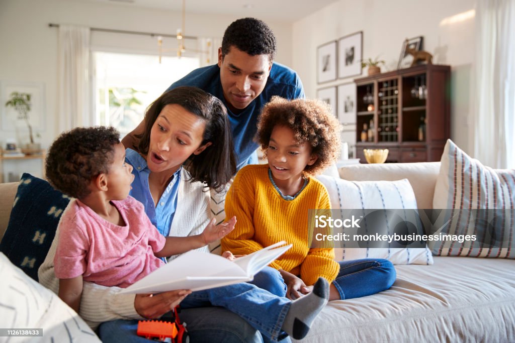 Young mother sitting on sofa in the living room with her daughter beside her and toddler son on her knee, father standing behind them, close up Family Stock Photo