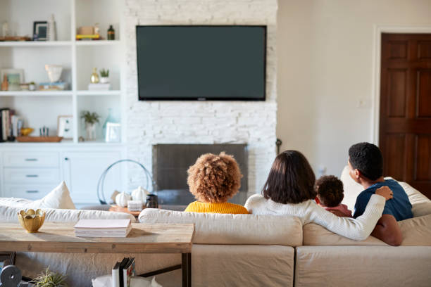 vue arrière du jeune famille assise sur le canapé et regardent la télévision ensemble dans leur salon - regarder la télévision photos et images de collection