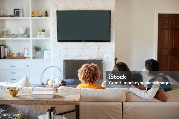 Photo libre de droit de Vue Arrière Du Jeune Famille Assise Sur Le Canapé Et Regardent La Télévision Ensemble Dans Leur Salon banque d'images et plus d'images libres de droit de Famille