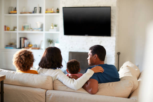 vue arrière du jeune famille assise sur le canapé et regarder la télévision ensemble dans leur salon, gros plan - arm around caucasian latin american and hispanic ethnicity child photos et images de collection