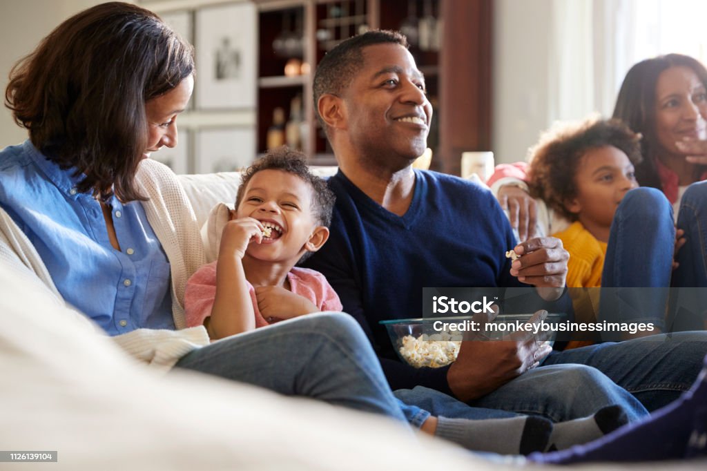Three generation family family sitting on the sofa in living room, watching TV and eating popcorn, selective focus Family Stock Photo