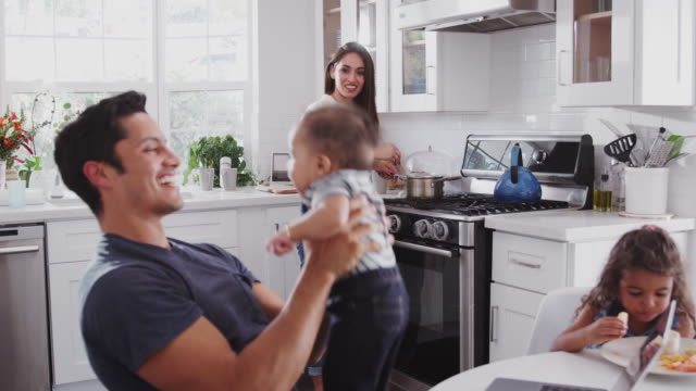 Hispanic family talking in their kitchen, mum cooking at hob, dad lifting baby in the air, close up