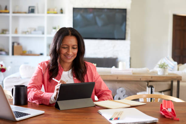 istock Smiling middle aged woman sitting at a table drawing on a tablet computer with a stylus, front view 1126141026