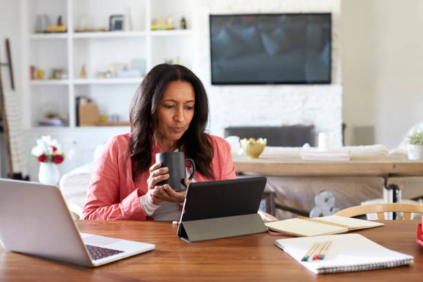 médio com idade mulher sentada em uma leitura de tabela usando um computador tablet, segurando uma taça, vista frontal - equipamento de telecomunicação - fotografias e filmes do acervo