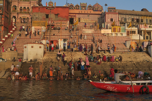 Varanasi, Uttar Pradesh India - March 27, 2017, Religious water ritual: Pilgrims and other Hindu believers are bathing in the Ganges river. Some have taken their clothes off, some are bathing in their shirts. In the background: Colourful red and white houses and the ghat stairs. In the foreground: Red boat with man rowing.