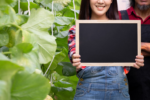 Close-up of beautiful smiling woman holding an empty blank black board inside green natural farm - with copy space to insert text