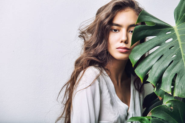 jeune fille belle modèle asiatique brune aux cheveux longs qui pose en studio avec une plante tropicale sur fond isolé - white green indoors studio shot photos et images de collection