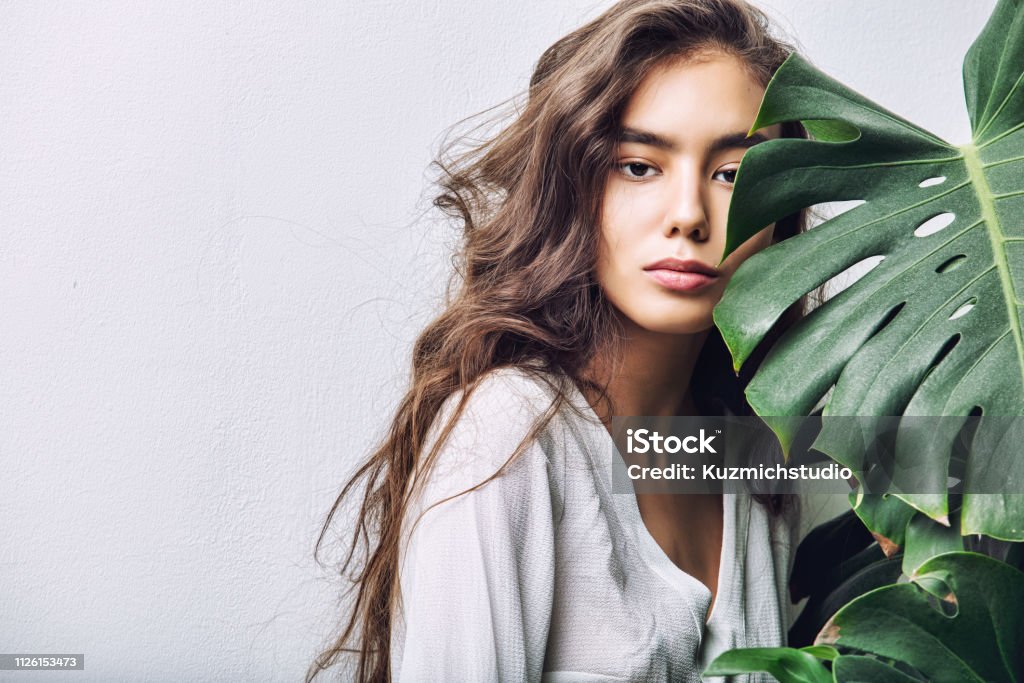 Young beautiful girl model Asian brunette with long hair posing in Studio with tropical plant on isolated background Beauty Stock Photo