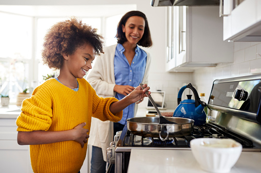 Pre-teen girl standing at hob in the kitchen using spatula and frying pan, preparing food with her mother, side view