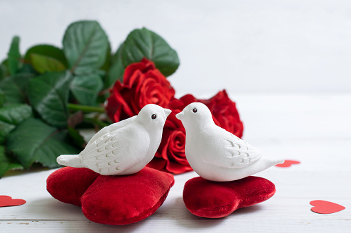 Home interior decor in honor of Valentine's Day with porcelain figurines of white kissing pigeons birds on velvet red decorative heart on background of roses bouquet