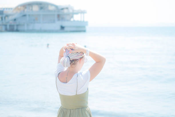 Durres, Albania, August 21, 2018: Young woman with multi-colored dreadlocks in a light cotton sundress stands on the beach at the water's edge and looks at the sea. View from the back