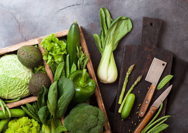 istock Assorted green toned raw organic vegetables in wooden box on dark background. Avocado, cabbage, cauliflower and cucumber with trimmed and mung beans and chopping board with knife. 1126183219