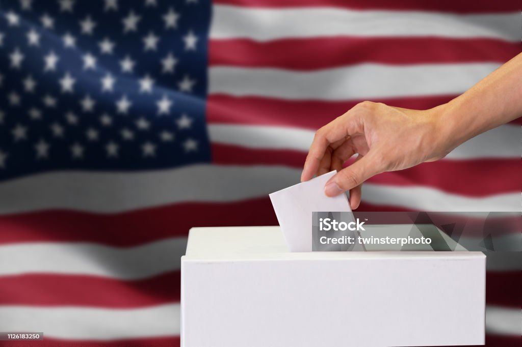 Close-up of man casting and inserting a vote and choosing and making a decision what he wants in polling box with United States flag blended in background. Close-up of man casting and inserting a vote and choosing and making a decision what he wants in polling box with United States flag blended in background.. Election Stock Photo
