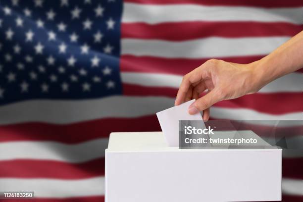 Closeup Of Man Casting And Inserting A Vote And Choosing And Making A Decision What He Wants In Polling Box With United States Flag Blended In Background Stock Photo - Download Image Now