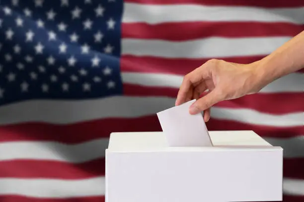 Photo of Close-up of man casting and inserting a vote and choosing and making a decision what he wants in polling box with United States flag blended in background.