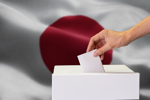 Close-up of human hand casting and inserting a vote and choosing and making a decision what he wants in polling box with Japan flag blended in background