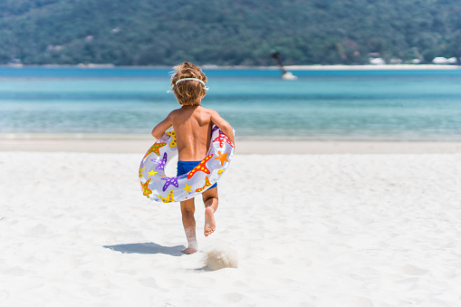 Back view of little boy with swimming tube running towards the sea in summer day. Copy space.