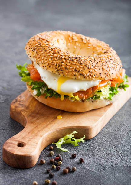 istock Fresh healthy bagel sandwich with salmon, ricotta and soft egg on vintage chopping board on stone kitchen table background. Healthy diet food. 1126192607