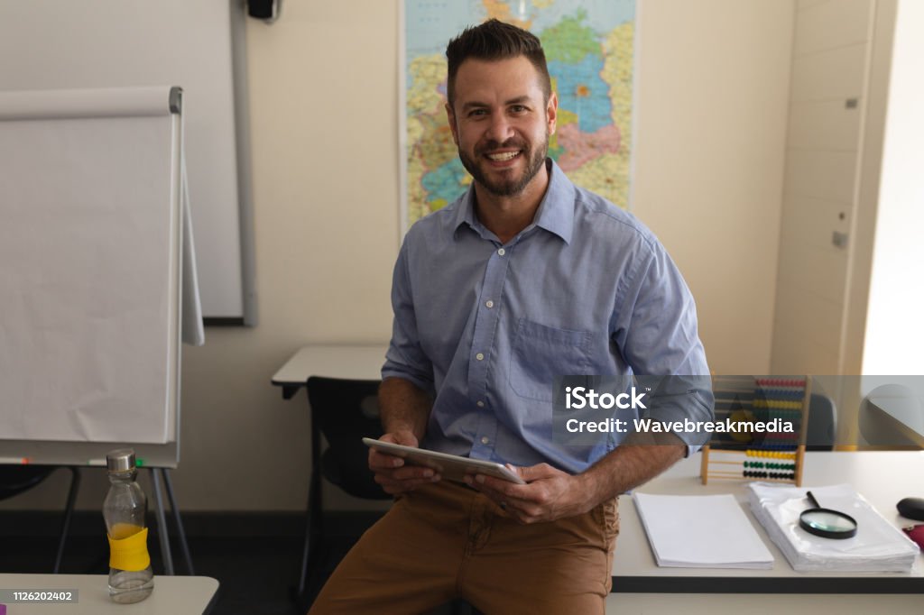 School teacher looking at camera and sitting on desk in classroom Happy school teacher with digital tablet looking at camera and sitting on desk in classroom Teacher Stock Photo