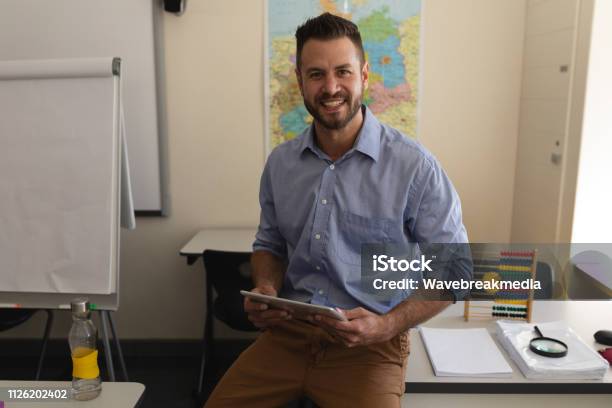 School Teacher Looking At Camera And Sitting On Desk In Classroom Stock Photo - Download Image Now