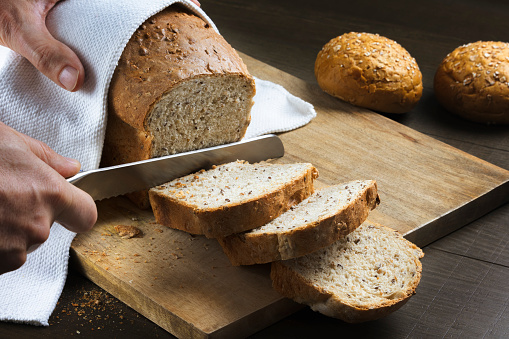 Whole grain bread put on rustic kitchen wood plate. Hands cutting bread with a serrated knife . Fresh bread on table close-up. copy space