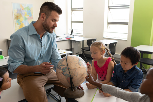 Side view of male teacher teaching his kids about geography by using globe in classroom of elementary school