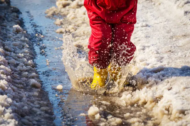 Kid legs in yellow rainboots jumping in the ice puddle with melting snow at sunny spring day, outdoors