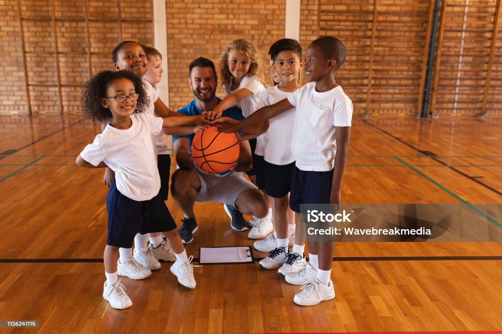 Schoolkids and basketball coach forming hand stack and looking at camera in basketball court Happy schoolkids and basketball coach forming hand stack and looking at camera in basketball court Child Stock Photo