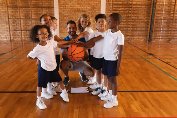 Happy schoolkids and basketball coach forming hand stack and looking at camera in basketball court