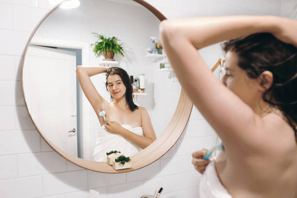 młoda atrakcyjna kobieta w białym ręczniku golenia pach, patrząc w lustrze w stylowej łazience. pielęgnacja skóry i ciała. koncepcja depilacji. kobieta po goleniu pod prysznicem z maszynką do golenia - shaving female shaving cream razor zdjęcia i obrazy z banku zdjęć