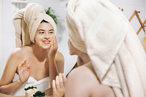 Concepto de cuidado de la piel. Mujer joven feliz en toalla haciendo masaje facial con exfoliante de la cara orgánica y mirando en el espejo en el cuarto de baño elegante. Chica aplicar la crema exfoliante, peeling y limpieza de piel photo