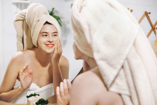 Skin Care concept. Young happy woman in towel making facial massage with organic face scrub and looking at mirror in stylish bathroom. Girl applying scrub cream, peeling and cleaning skin stock photo