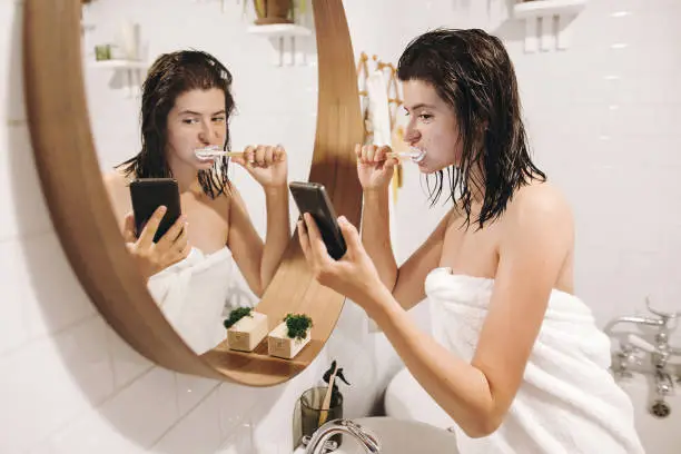 Photo of Social media affect. Young happy woman in white towel brushing teeth and looking at phone screen in stylish bathroom at round mirror. Slim sexy woman daily routine after shower.