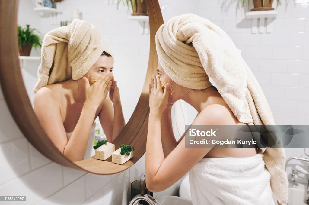 Jeune femme heureuse en serviette appliquant le gommage visage bio et en regardant un miroir rond dans l’élégante salle de bains. Fille faisant un massage du visage, gommage et nettoyage de peau sur le visage. Soins de la peau et l’hygiène - Photo de Exfoliation libre de droits