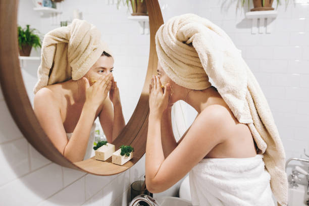 young happy woman in towel applying organic face scrub and looking at round mirror in stylish bathroom. girl making facial massage, peeling and cleaning skin on face. skin care and hygiene - beauty treatment spa treatment health spa human face imagens e fotografias de stock